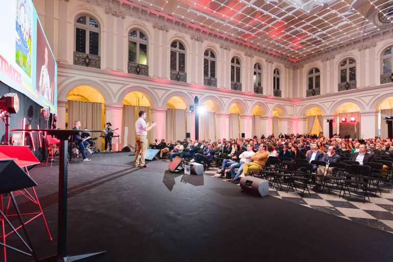 Un orateur se tient sur scène et s'adresse à un public assis dans une grande salle aux fenêtres cintrées et au sol en damier. Le public attentif écoute attentivement la French Tech Night 2023. La salle est décorée d'un éclairage tamisé et un grand écran diffuse des images à côté de la scène.