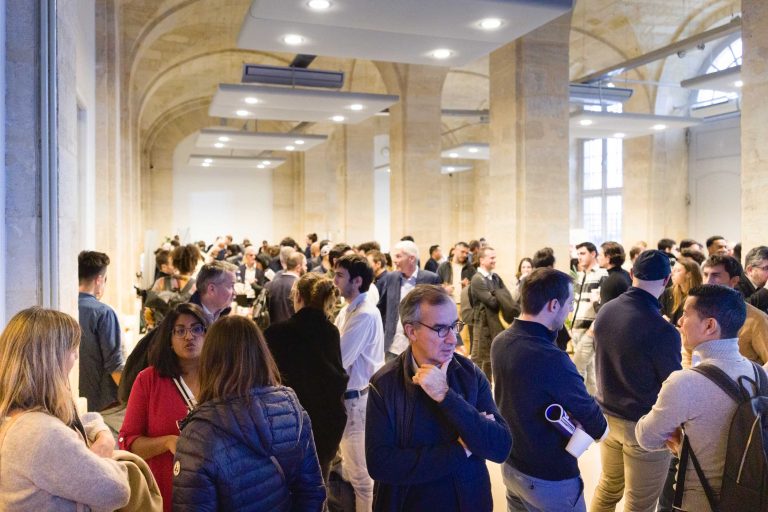 Une salle spacieuse et bien éclairée, avec de hauts plafonds et des arches en pierre, est remplie de nombreuses personnes engagées dans des conversations. La foule, habillée de tenues décontractées ou professionnelles, semble diversifiée en termes d'âge et de sexe. Certains individus tiennent des boissons ou des documents, suggérant un événement professionnel ou de réseautage comme la French Tech Night 2023.