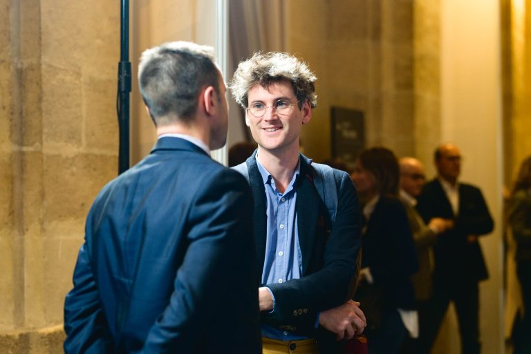 Deux hommes discutent à la French Tech Night 2023. L'homme de gauche, aux cheveux courts, regarde ailleurs, vêtu d'un costume sombre. L'homme de droite, aux cheveux bouclés, portant des lunettes et une chemise bleue sous un blazer, sourit chaleureusement. Un arrière-plan flou révèle d'autres participants dans une salle aux murs de pierre à la lumière chaleureuse.