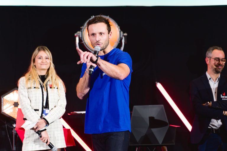 Lors de la French Tech Night 2023, un homme en chemise bleue parle dans un micro sur scène en faisant des gestes de la main. À sa gauche, une femme en veste blanche sourit, tenant des papiers. À sa droite, un homme en blazer sombre regarde en souriant. Des lumières vives et du matériel de scène sont visibles en arrière-plan.