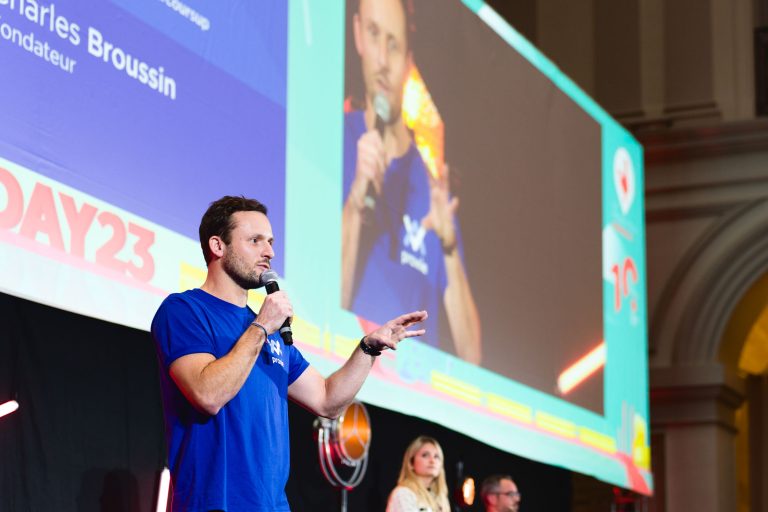 Un homme tenant un micro s'exprime sur scène lors de la French Tech Night 2023. Il porte un t-shirt bleu et se tient debout devant un grand écran qui affiche son image et son nom. Deux autres individus sont assis derrière lui sur scène.