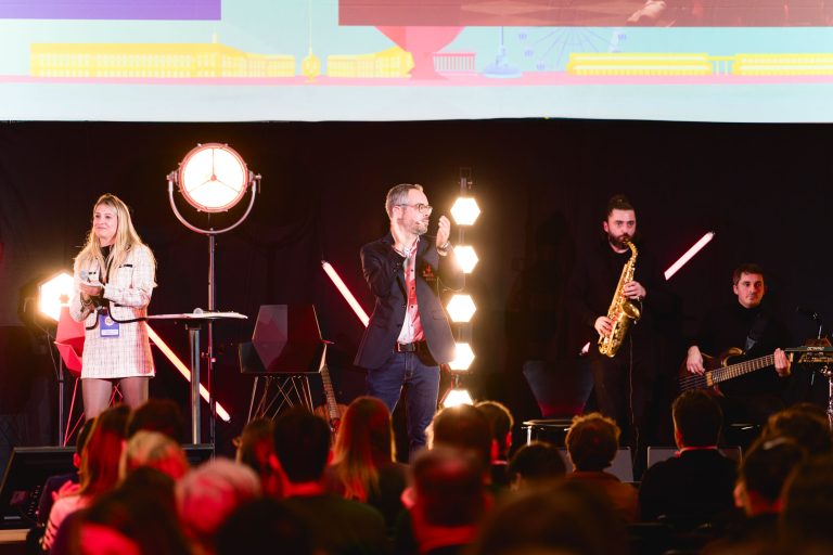 Lors de la French Tech Night 2023, un groupe de quatre personnes est sur scène. Une femme tenant un micro et souriante se tient à gauche, tandis qu'un homme à côté d'elle applaudit. À droite, un homme joue du saxophone et un autre de la guitare. Des membres du public sont présents, avec des lumières de scène et un écran visible en arrière-plan.