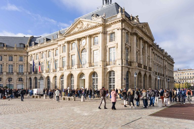 Une foule animée se rassemble devant un bâtiment historique néoclassique de couleur crème, avec des fenêtres cintrées et des colonnes. Des drapeaux ornent la façade du bâtiment alors que la French Tech Night 2023 démarre. Le ciel est partiellement nuageux, ce qui ajoute à l'atmosphère animée de la place pavée.