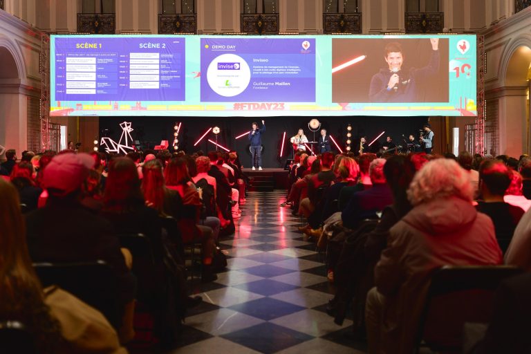 Un large public est assis en rangs face à une scène avec deux écrans et un orateur tenant un micro au centre. L'événement, la French Tech Night 2023, se déroule dans une grande salle avec de hautes arches et des moulures décoratives. Le public regarde attentivement l'écran, montrant les détails de l'événement et un personnage parlant. #FTDAY23.