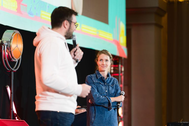 Deux personnes se tiennent sur scène avec des micros lors de la French Tech Night 2023. La personne de gauche, vêtue d'un sweat à capuche blanc, parle, tandis que celle de droite, vêtue d'une chemise en jean bleu, écoute attentivement. L'arrière-plan présente une projection colorée sur un écran, avec un éclairage de scène positionné à gauche.