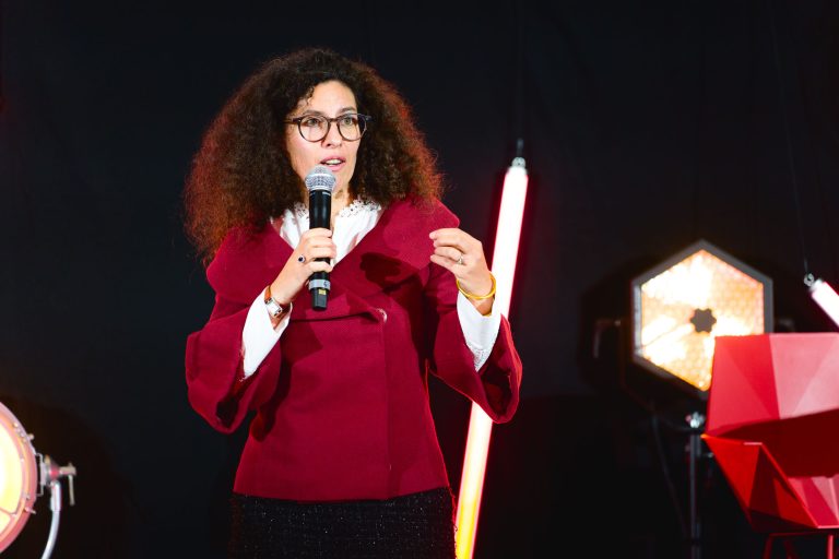 Une femme aux cheveux bouclés et aux lunettes, vêtue d'une veste rouge, parle dans un micro. Elle fait des gestes de la main gauche. L'arrière-plan est sombre avec des lumières allumées : une lampe de studio hexagonale à droite et des tubes lumineux rouges en arrière-plan, préparant le terrain pour la French Tech Night 2023.