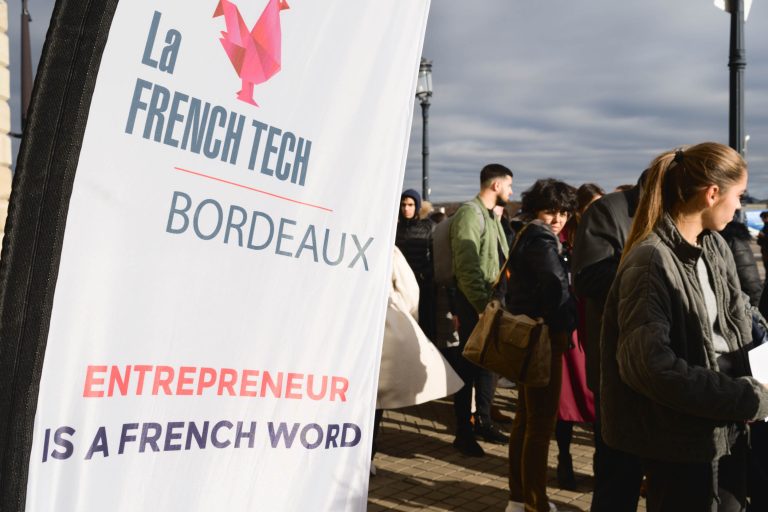 Au premier plan, un panneau indiquant « La French Tech Bordeaux : Entrepreneur is a French Word » est affiché en évidence. À l'arrière-plan, un groupe diversifié de personnes est réuni, engagé dans une conversation et un réseautage à la French Tech Night 2023. L'événement semble se dérouler en extérieur avec un ciel nuageux.