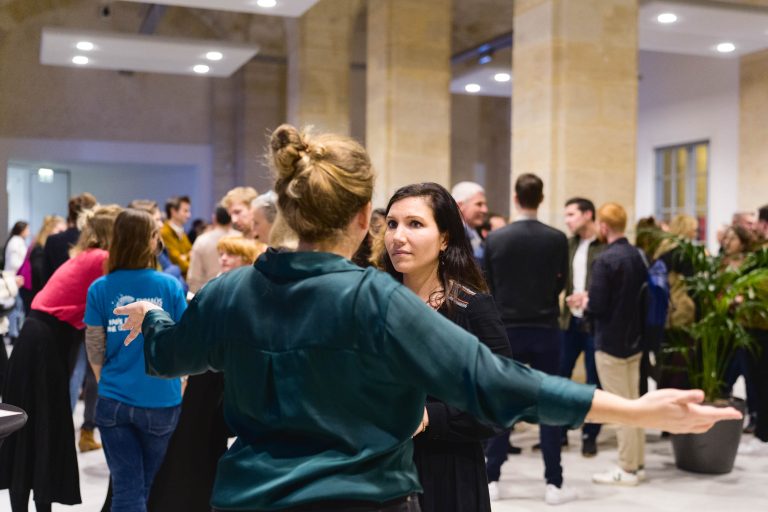 Une soirée French Tech 2023 animée dans un espace lumineux et moderne aux murs beiges. Au premier plan, une femme aux bras ouverts, dos à la caméra, parle à une autre femme aux longs cheveux bruns. De nombreuses personnes discutent en arrière-plan. Une personne en chemise bleue est visible à gauche.