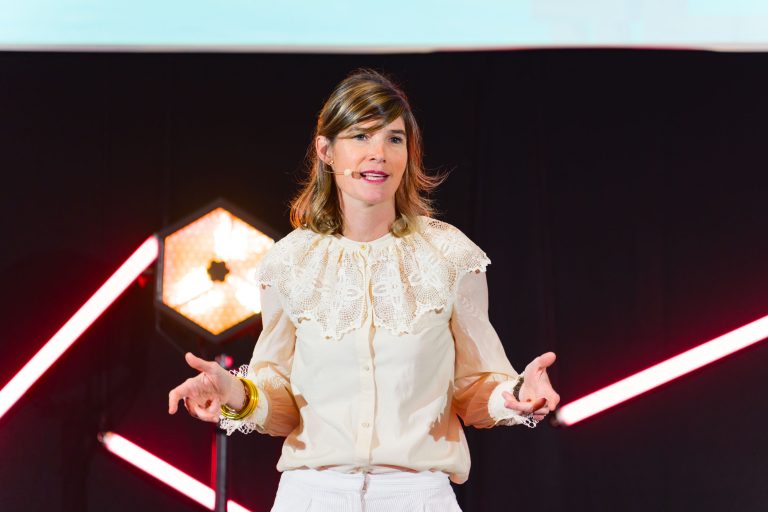 Une femme aux cheveux châtain clair donne une conférence sur scène à la French Tech Night 2023. Elle porte une blouse couleur crème avec des détails en dentelle sur le col et un pantalon blanc. Elle a un micro et fait des gestes avec ses mains. L'arrière-plan présente des lumières diagonales rouges et une lumière hexagonale.