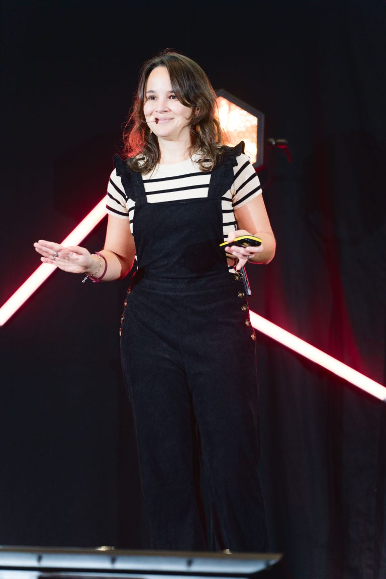 Lors de la French Tech Night 2023, une femme se tient sur scène en train de parler, vêtue d'une chemise rayée et d'une salopette noire. Elle a les cheveux longs et une expression confiante, une main faisant un geste et l'autre tenant un objet. Des lumières de scène rouges et blanches et un fond sombre soulignent sa présence.