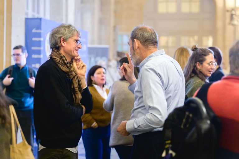 Un groupe de personnes se rassemble dans un espace intérieur spacieux et bien éclairé pour discuter. Deux hommes discutent au premier plan, l'un portant un foulard et des lunettes, l'autre une chemise bleue. L'ambiance animée de la French Tech Night 2023 fait que d'autres personnes en arrière-plan discutent également.