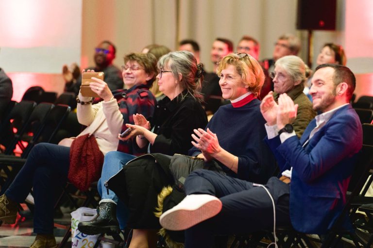 Lors de la French Tech Night 2023, un groupe diversifié de personnes est assis sur des rangées de chaises, participant attentivement à l'événement en salle avec un éclairage d'accentuation rouge. Plusieurs individus applaudissent, certains sourient et une personne capture l'instant avec un téléphone. La scène transmet un public positif et engagé.