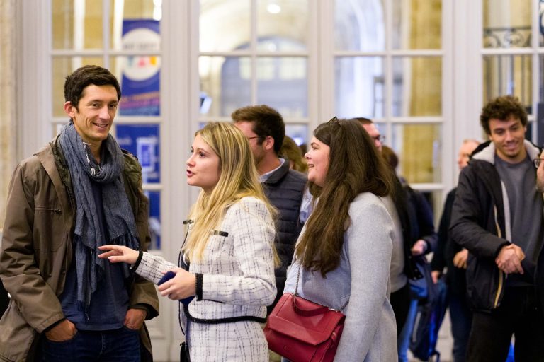 Un groupe de personnes est réuni à l'intérieur de la French Tech Night 2023, discutant et souriant. Le décor semble être une salle bien éclairée avec de grandes fenêtres et des portes en arrière-plan. Une femme en blouse blanche fait des gestes tout en parlant à un homme portant un foulard. D'autres personnes à proximité écoutent et rient.