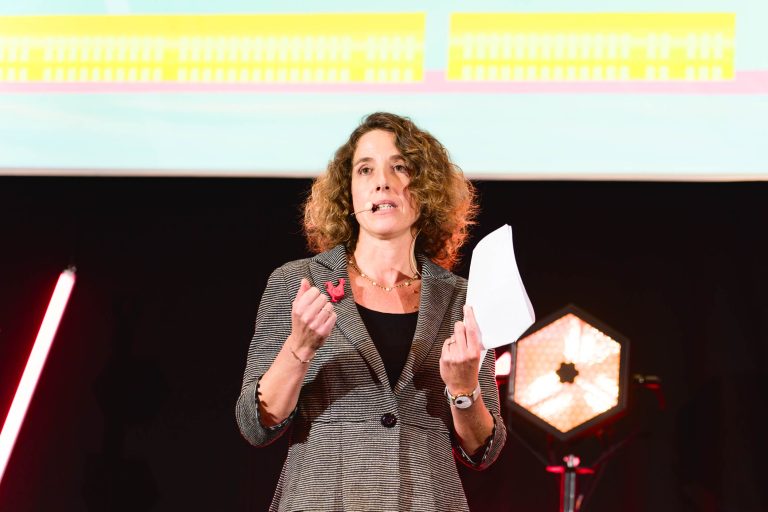 Lors de la French Tech Night 2023, une femme aux cheveux bouclés s'exprime avec passion sur scène, tenant un morceau de papier dans une main et faisant des gestes de l'autre. Elle porte un blazer gris et un micro-casque. L'arrière-plan comprend un écran lumineux affichant des graphiques multicolores et des lumières de scène, dont l'une est visible derrière elle.