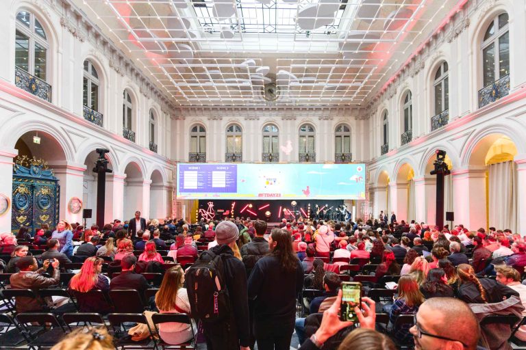 Un large public est réuni dans une grande salle au haut plafond vitré et aux arches ornementées, face à une scène sur laquelle est projeté un grand écran diffusant une présentation. À la French Tech Night 2023, les gens sont assis et debout, certains prennent des photos avec leur téléphone. L'ambiance est animée et animée.