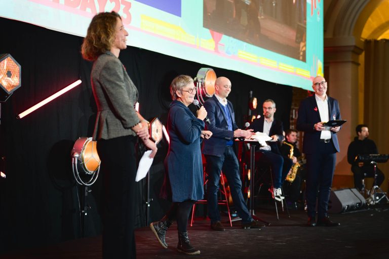 Un groupe de cinq personnes est sur scène pour participer à la French Tech Night 2023. Une femme en robe bleue parle dans un micro, tandis que trois hommes et une femme se tiennent debout, tenant des papiers ou des tablettes. Des lumières de scène et un grand écran affichant des graphiques et du texte remplissent l'arrière-plan.