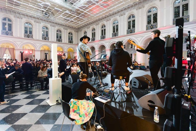 Un groupe se produit sur scène dans une grande salle élégante à l'architecture classique et aux hauts plafonds en verre pendant la French Tech Night 2023. Les membres du groupe jouent d'un instrument et le public est assis, regardant attentivement. La salle est bien éclairée et les gens sont rassemblés, ce qui indique un événement en direct ou un concert.