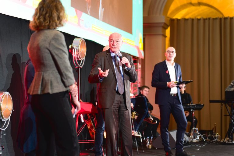 Lors de la French Tech Night 2023, un homme d'un certain âge parle dans un micro sur scène, en faisant des gestes de la main gauche. Entouré d'une femme en blazer gris et d'un homme à lunettes tenant une tablette, ils sont placés devant un grand écran et un éclairage d'événement. D'autres participants et équipements sont visibles.