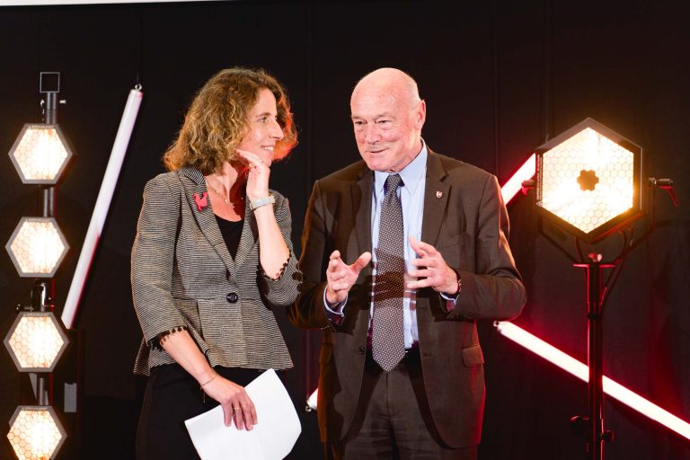 Lors de la French Tech Night 2023, une femme aux cheveux bouclés et un homme en costume discutent sur scène, illuminés par des lumières géométriques en arrière-plan. La femme tient un document, tandis que l'homme fait des gestes avec ses mains, créant une atmosphère de discussion animée. Tous deux semblent concentrés et animés.