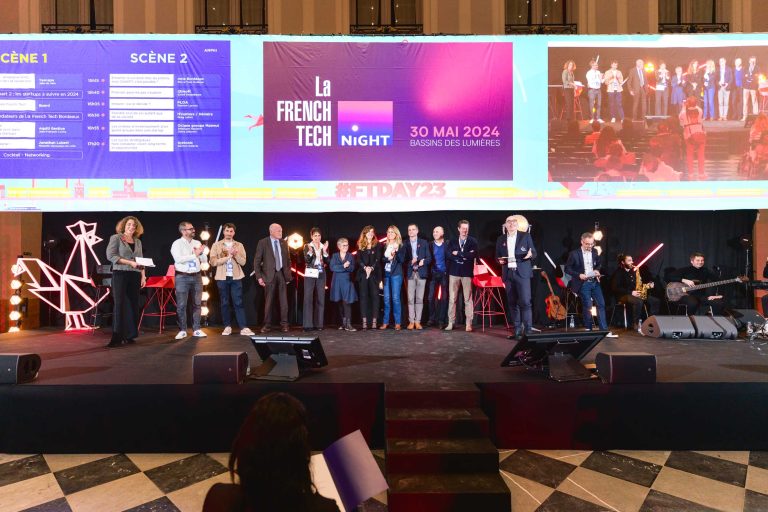 Un groupe diversifié de personnes se tient sur scène lors de l'événement French Tech Night 2023. L'arrière-plan présente deux grands écrans affichant les détails de l'événement : « 30 mai 2024 » et « Bassin des Lumières ». Un éclairage très lumineux et une installation avec des instruments de musique à droite. Un public engagé est visible au premier plan.