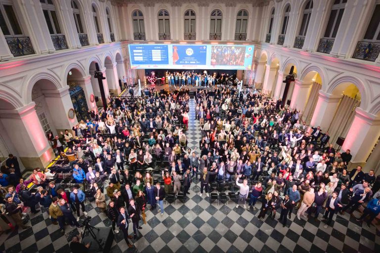 Un grand rassemblement de personnes se tient dans une grande salle au sol en damier, aux colonnes arquées et au balcon à l'étage. En arrière-plan, un grand écran affiche des informations sur la French Tech Night 2023. Une scène et des banderoles plantent le décor tandis que les participants semi-formels se tournent vers l'avant, certains faisant signe de la main ou applaudissant.