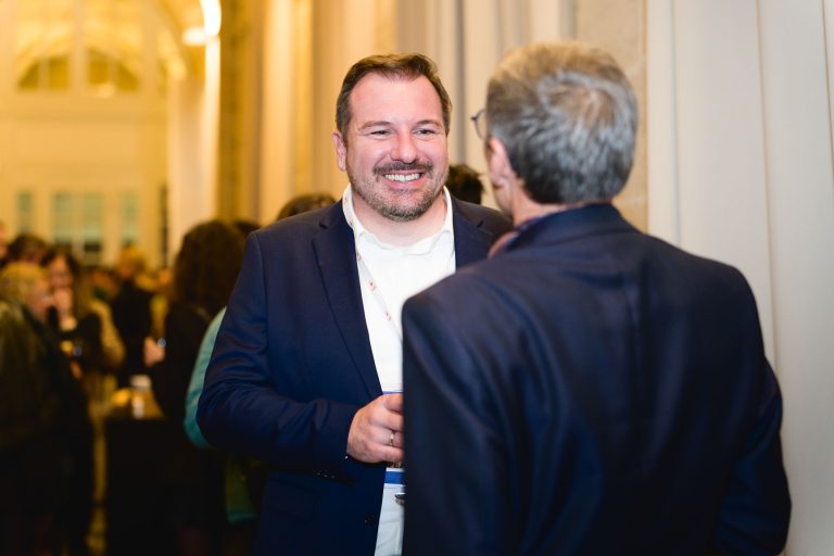 Un homme en costume sombre et chemise blanche sourit et tient un verre tout en discutant avec un autre homme, également vêtu d'un costume sombre. L'arrière-plan révèle l'ambiance animée de la French Tech Night 2023, avec un éclairage chaleureux et de nombreux autres participants floutés dans le décor.