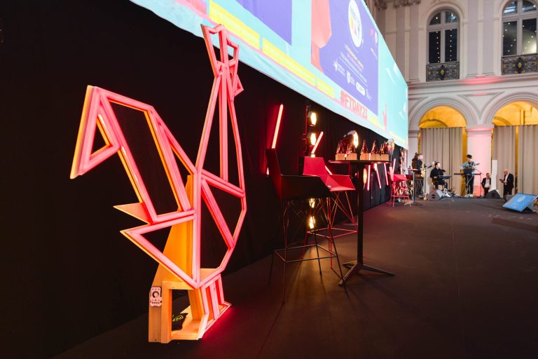 Une scène événementielle moderne pour la French Tech Night 2023 présente à gauche un grand coq géométrique illuminé. À droite, une rangée de chaises hautes et de tables avec éclairage d'ambiance sur fond noir. En arrière-plan, un grand écran diffuse des visuels colorés.