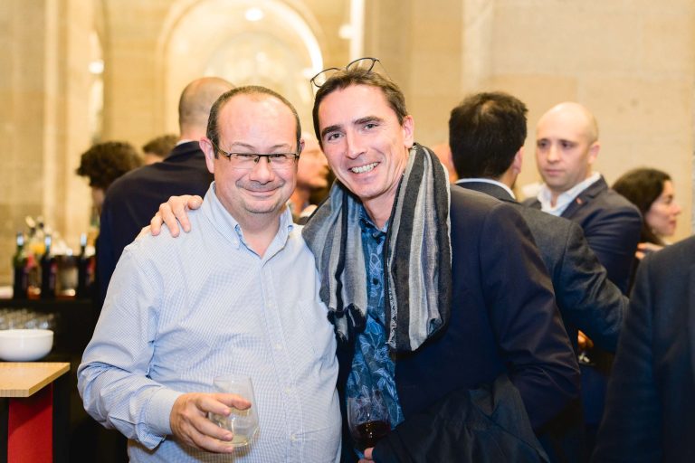 Lors de la French Tech Night 2023, deux hommes se tiennent côte à côte lors d'une réunion sociale. L'homme de gauche, portant des lunettes et une chemise bleu clair, tient un verre. L'homme de droite a son bras autour de ses épaules, vêtu d'un blazer sombre et d'une écharpe, tenant également un verre. Les gens se mêlent en arrière-plan.