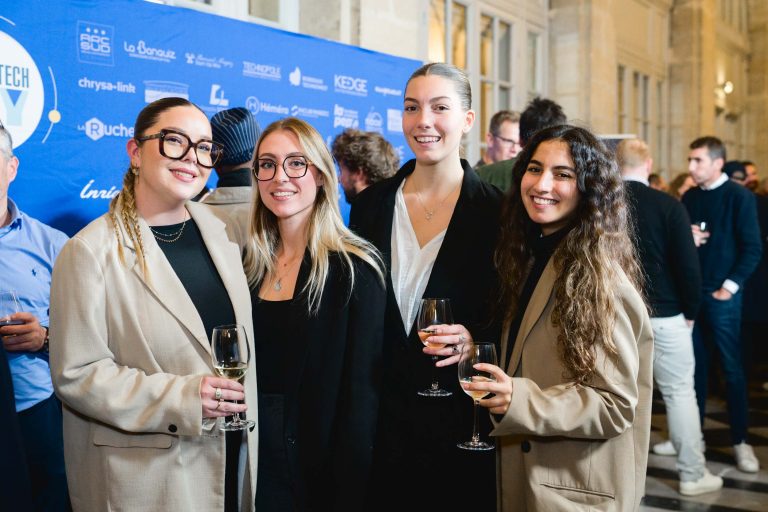 Quatre femmes se tiennent debout, tenant des verres à vin à la French Tech Night 2023. Elles sont habillées en tenue professionnelle avec des blazers beiges, des chemises blanches et des hauts noirs. Derrière elles, un fond bleu de sponsor. D'autres participants se mêlent en arrière-plan, créant une atmosphère sophistiquée et professionnelle.