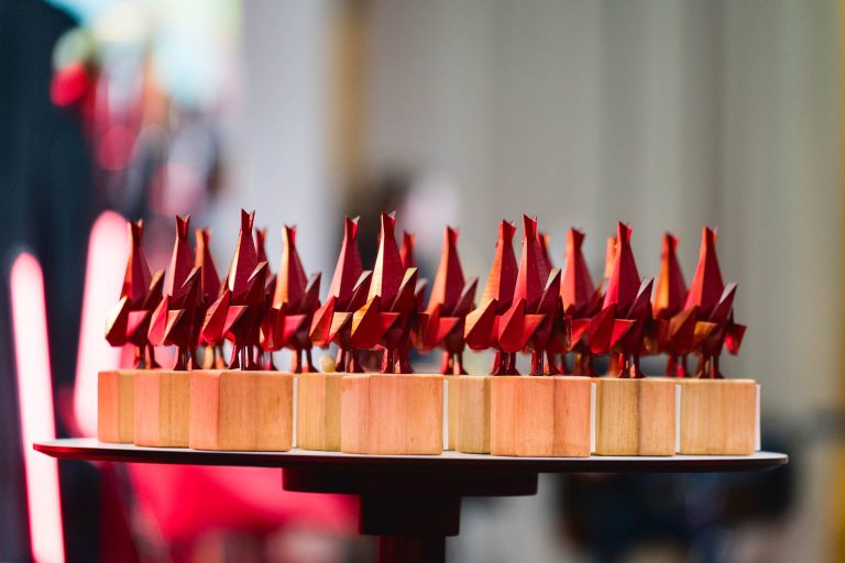 Plusieurs sculptures d'oiseaux rouges identiques en origami sont soigneusement disposées sur des blocs de bois individuels. Ces blocs sont placés sur une plateforme ronde noire, avec un arrière-plan flou composé de divers objets et couleurs indistincts, suggérant un cadre formel ou artistique à la French Tech Night 2023.