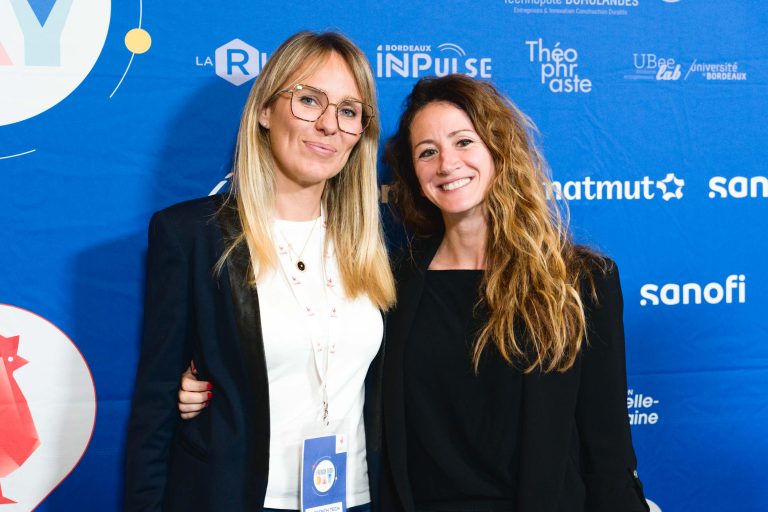 Deux femmes se tiennent devant un fond bleu sur lequel figurent des logos tels que « Sanofi », « Matmut » et « UB Sanctis », entre autres, lors de la French Tech Night 2023. La femme de gauche a les cheveux blonds, porte des lunettes et un blazer noir. La femme de droite a les cheveux longs et ondulés et porte une veste noire. Toutes deux sourient et se prennent dans les bras