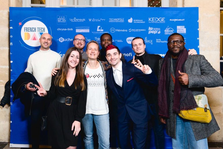 Un groupe de huit personnes se tient debout devant un fond photographique bleu avec divers logos et les mots « French Tech Night 2023 ». Ils sourient et posent, certains faisant des signes de paix et des gestes de pouce levé. Le groupe est habillé d'un mélange de tenues décontractées et semi-formelles.