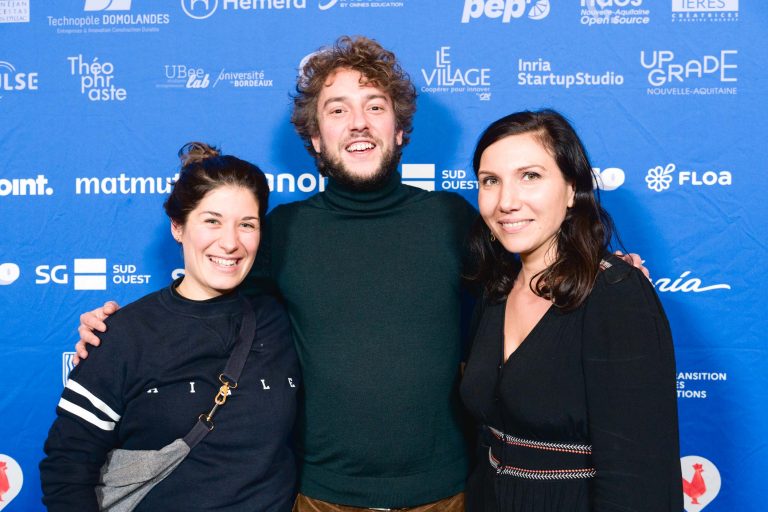 Trois individus se tiennent rapprochés, souriant devant un fond bleu vif comportant divers logos blancs de sponsors et d'organisations de la French Tech Night 2023. La personne de gauche porte un sweat-shirt foncé, la personne du milieu porte un col roulé foncé et la personne de droite porte un haut noir.