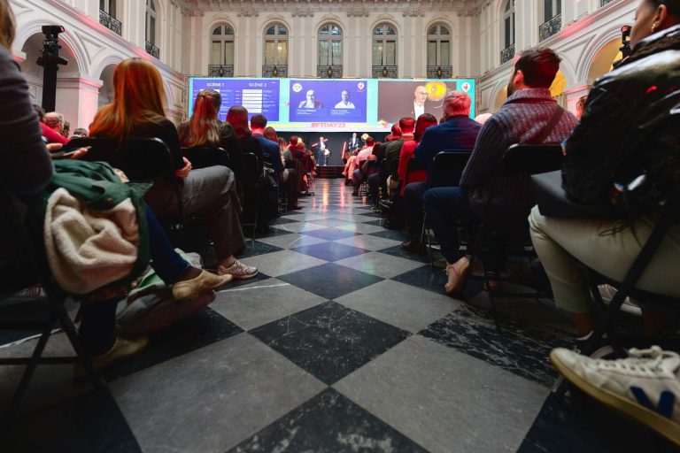 Un public hétéroclite est assis sur des rangées de chaises dans une grande salle aux hauts plafonds. Ils regardent une présentation diffusée sur un grand écran à l'avant de la French Tech Night 2023. Le sol est orné d'un motif à carreaux noir et blanc, et plusieurs personnes à l'autre bout se tiennent derrière un podium.