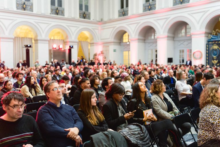 Un grand groupe de personnes est assis avec attention dans une grande salle aux murs blancs et aux fenêtres cintrées. De nombreuses personnes sont concentrées sur la scène, certaines utilisent leur téléphone ou discutent. La salle, qui accueille la French Tech Night 2023, est dotée d'un éclairage chaleureux et de piliers décorés, créant une atmosphère élégante et formelle.