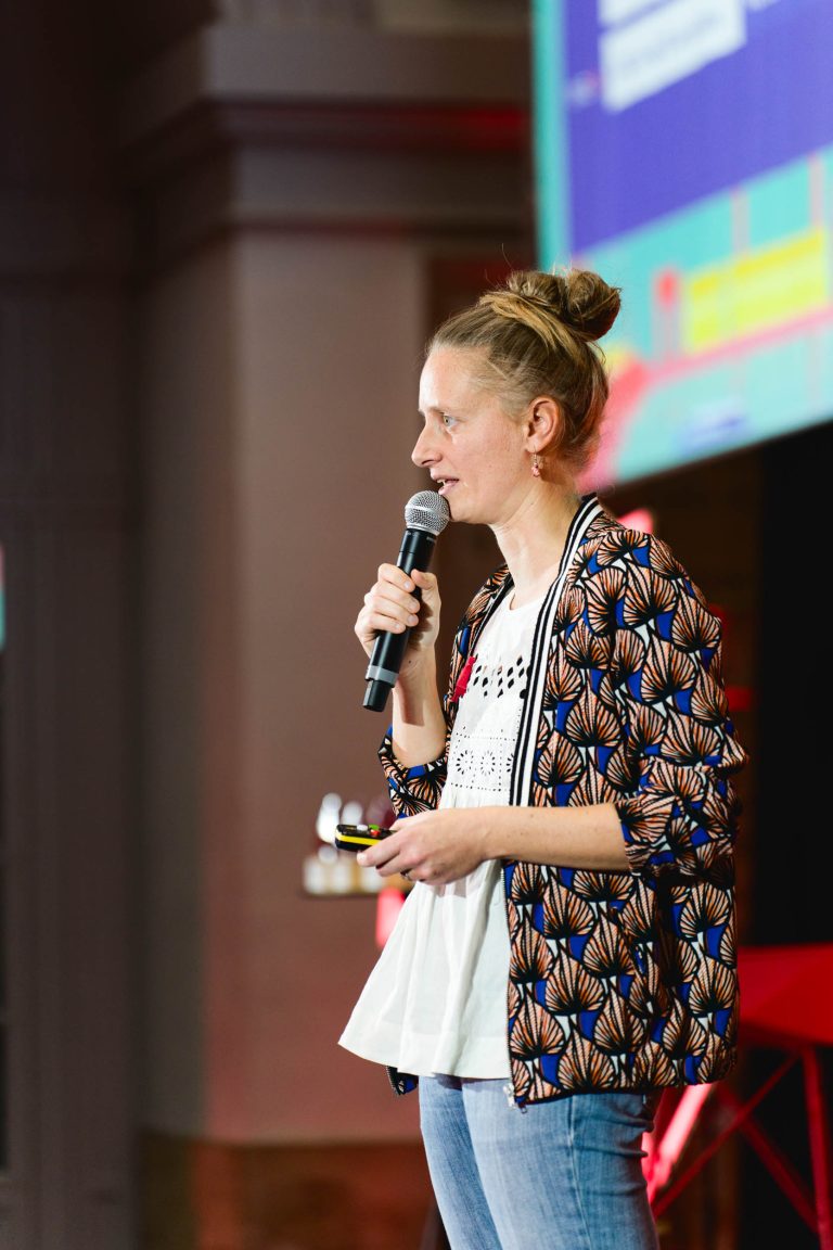 Lors de la French Tech Night 2023, une personne aux cheveux blonds coiffés en chignon parle dans un micro, tenant un petit objet dans l'autre main. Elle porte un blazer à motifs sur un t-shirt blanc et un jean. Elle présente son exposé devant un écran aux graphismes colorés. Le décor semble être celui d'un événement ou d'une conférence en salle.