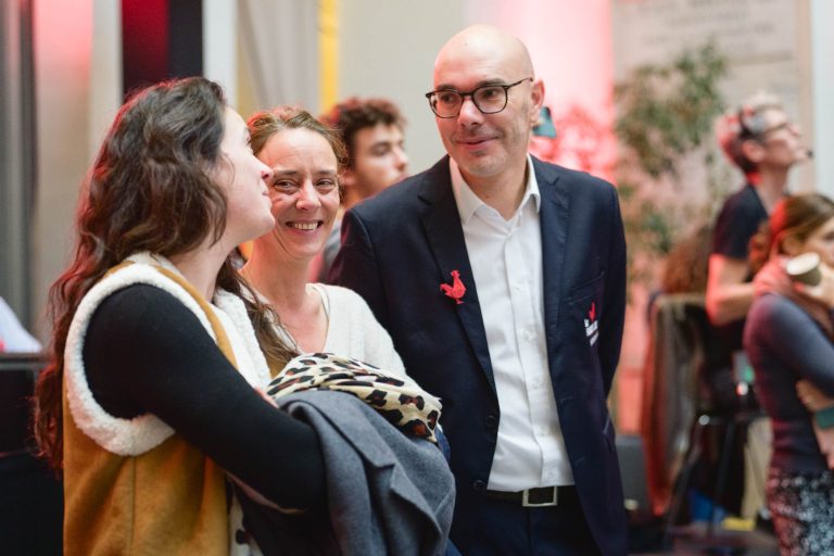 Trois personnes en tenue semi-formelle discutent dans l'enceinte de la French Tech Night 2023. L'homme de droite, en costume et lunettes, regarde attentivement les deux femmes de gauche. La femme du milieu, souriante, fait face à la femme en gilet et écharpe, tenant une veste. D'autres sont floues en arrière-plan.