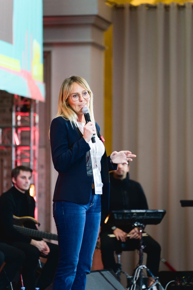 Une femme aux cheveux blonds et aux lunettes parle dans un micro sur scène lors de la French Tech Night 2023. Elle porte un blazer foncé, une chemise blanche et un jean. Derrière elle, un homme est assis avec une guitare et un autre individu est près d'un équipement musical. Un grand écran et un éclairage de scène sont visibles en arrière-plan.