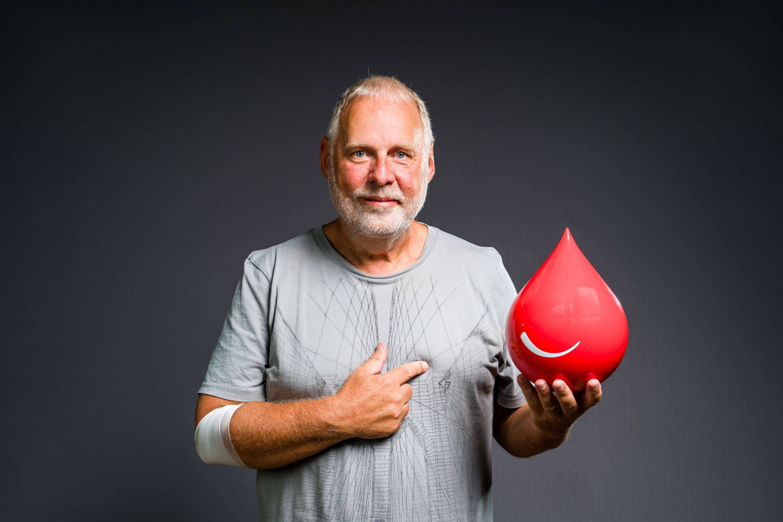 Un homme âgé avec une barbe poivre et sel et une chemise gris clair sourit en désignant une grosse goutte rouge brillante avec un smiley blanc dessus. Son bras droit a un bandage blanc près du coude. Le fond est d'un gris uni, rappelant les portraits professionnels à Bordeaux.