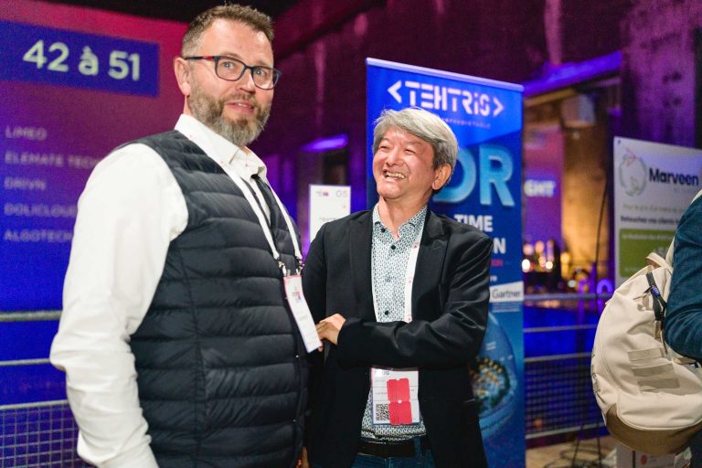 Deux hommes sont debout et sourient à la French Tech Night 2024 avec des banderoles en arrière-plan. L'homme de gauche porte des lunettes et une barbe, porte une chemise blanche et un gilet noir. L'homme de droite a les cheveux gris, porte une chemise à motifs et un blazer noir. Des personnes et des affichages d'événements sont visibles derrière eux.