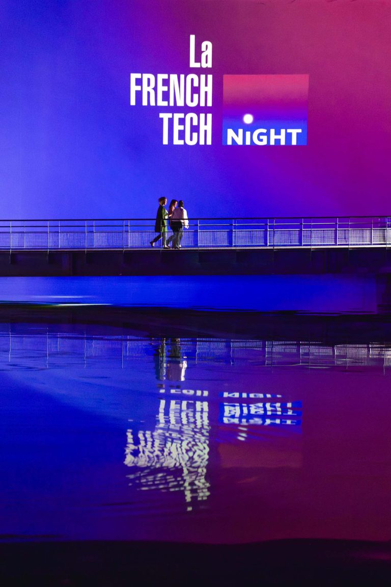 Un couple se tient debout sur un pont au-dessus d'une eau calme, qui reflète les mots « French Tech Night 2024 » projetés sur le mur devant eux. La projection présente un texte blanc en gras et un logo en dégradé rouge et bleu, créant un arrière-plan illuminé avec un mélange de teintes violettes, bleues et rouges.