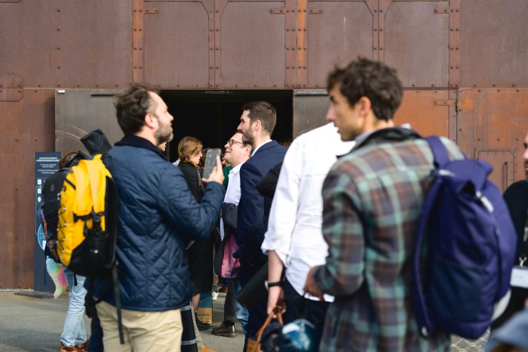 Un groupe de personnes, principalement des hommes, se tient debout et discute près de l'entrée d'un bâtiment à la façade brun-rougeâtre et patinée par les intempéries. Une personne porte un sac à dos jaune, tandis qu'une autre porte une veste à carreaux. La scène, qui se déroule pendant la French Tech Night 2024, semble décontractée et sociale avec des personnes habillées en tenue semi-décontractée.