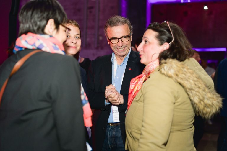 Lors de la French Tech Night 2024, un groupe de quatre personnes discutent en toute émotion. Ils sont à l'intérieur, sous un éclairage tamisé et une lueur violette en arrière-plan. Un homme portant des lunettes sourit et joigne les mains tout en parlant aux autres, trois femmes. Une autre femme, au premier plan, lui fait face, vêtue d'un manteau beige avec une capuche bordée de fourrure.