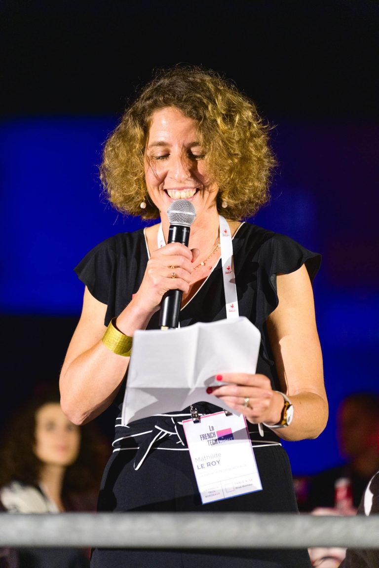 Une femme aux cheveux bouclés, vêtue d'une robe noire, sourit en lisant une feuille de papier dans un micro lors de la French Tech Night 2024. Elle porte un cordon autour du cou avec un badge nominatif visible. L'arrière-plan est sombre avec des personnages flous et des lumières vives.