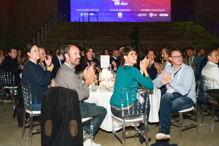 Un groupe de personnes est assis autour de tables lors de la French Tech Night 2024, applaudissant et regardant vers la scène. La salle est faiblement éclairée par un éclairage doux qui se concentre sur le public. La toile de fond comprend un escalier et un grand écran avec du texte en arrière-plan. Certains participants ont des boissons sur les tables.