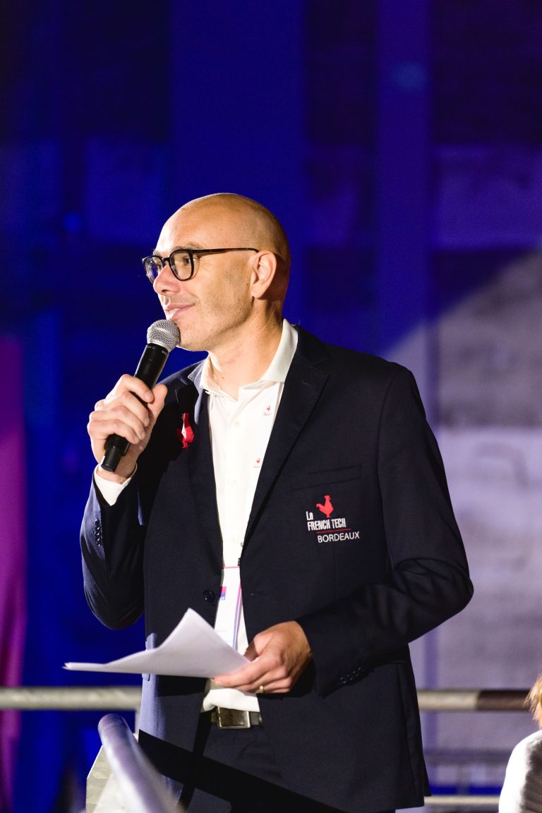 Un homme portant des lunettes, un blazer noir avec l'inscription « French Tech Bordeaux » brodée sur la poitrine et une chemise blanche tient un micro dans sa main droite et des papiers dans sa main gauche. S'adressant au micro sur fond violet à l'intérieur, il s'adresse à la foule lors de la French Tech Night 2024.