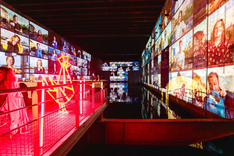 Une installation artistique dynamique et immersive à la French Tech Night 2024 comprend de nombreux écrans affichant différentes personnes, remplissant les murs de la salle. Une structure lumineuse au néon rouge se trouve sur une passerelle au-dessus d'une surface d'eau réfléchissante qui reflète les visuels illuminés. Une personne se tient debout à gauche, regardant l'exposition.