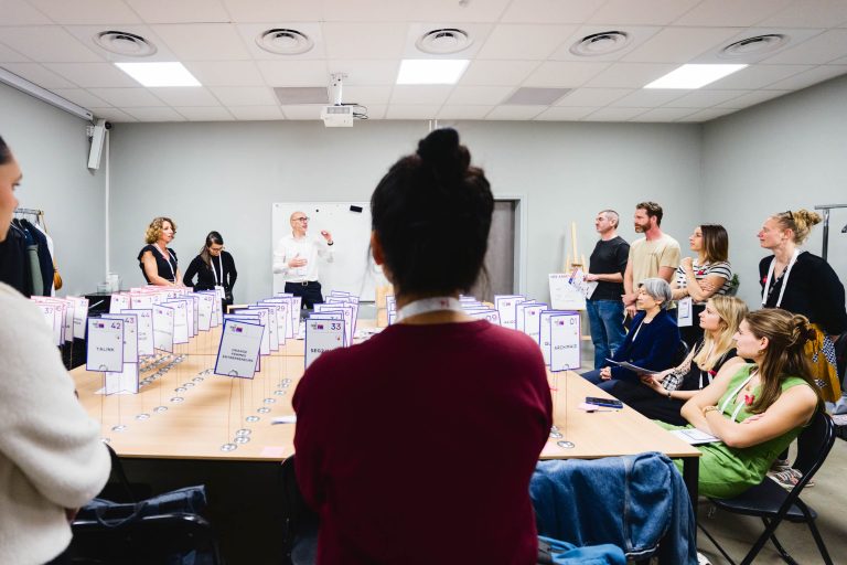 Lors de la French Tech Night 2024, un groupe d’individus se réunit dans une salle autour de deux tables disposées en U. Les tables sont couvertes de nombreux dossiers ou documents et de feutres numérotés. Une personne debout près d’un tableau blanc parle, tandis que les autres, assis et debout, écoutent attentivement.