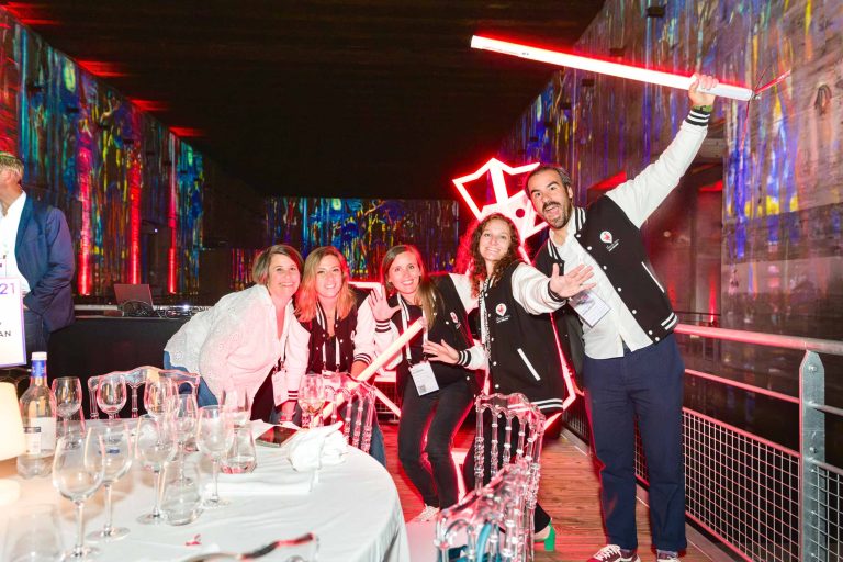 Un groupe de cinq personnes, trois femmes et deux hommes, posent joyeusement lors de l'événement haut en couleur de la French Tech Night 2024. Ils portent tous des vestes noires et blanches assorties et tiennent des bâtons rouges lumineux. Le décor est vibrant, avec des lumières multicolores projetées sur les murs. Divers objets en verre sont disposés sur une table au premier plan.