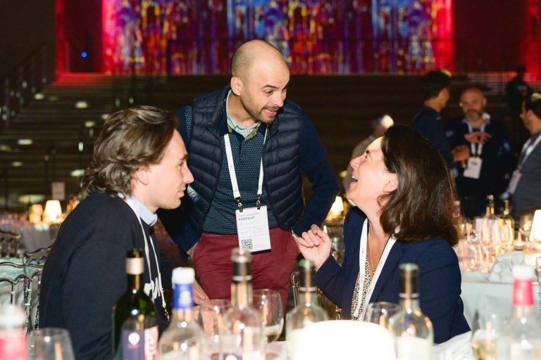 Trois personnes discutent avec enthousiasme autour d'une table dressée avec des bouteilles de vin et des verres dans un lieu élégant et à la lumière tamisée pendant la French Tech Night 2024. L'arrière-plan présente des œuvres d'art abstraites colorées et illuminées. La personne debout sourit, tandis que les deux personnes assises sont animées et souriantes.